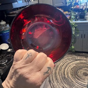 May include: A large, ruby-red glass bowl with a wide rim. The bowl is held in a hand with tattoos on the knuckles. The bowl's interior reflects light, creating a vibrant, glossy appearance. The bowl is likely for serving or display.