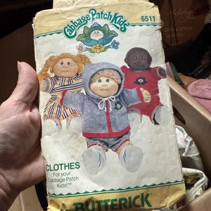 May include: A Butterick sewing pattern envelope for Cabbage Patch Kids clothes, model 6511. The packaging features illustrations of three dolls wearing various outfits, including a grey hooded jacket, a red shirt, and a striped dress, with green text.