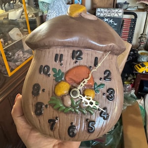 May include: A brown, acorn-shaped wall clock with a textured surface, featuring raised black numbers from 1 to 12. The center displays a painted design of orange and yellow mushrooms with green leaves, complemented by gold-colored clock hands.