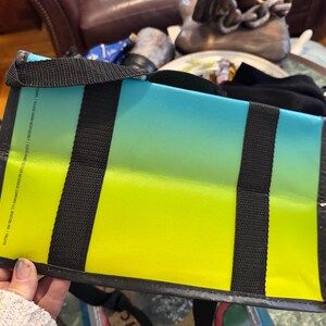 May include: A rectangular tote bag with a gradient design, transitioning from yellow to blue. The bag features black straps and a black trim. The bag is held by a person.