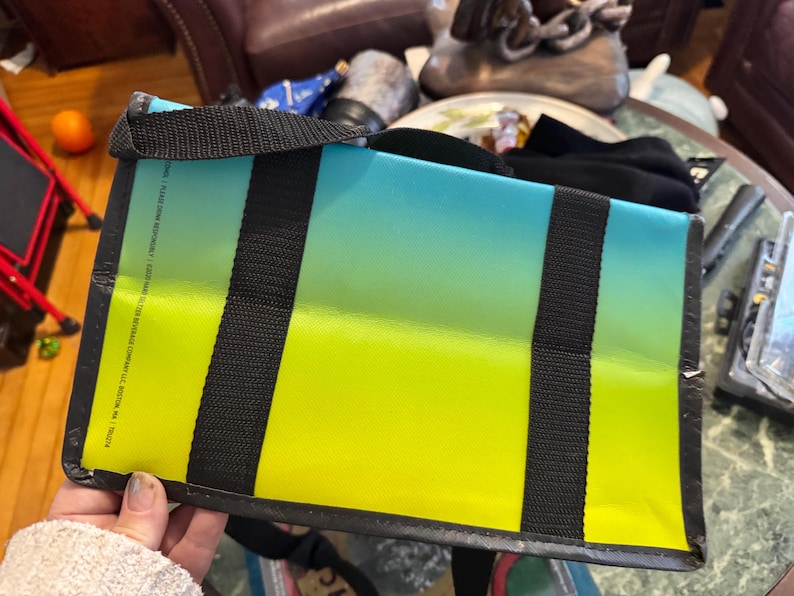 May include: A rectangular tote bag with a gradient design, transitioning from yellow to blue. The bag features black straps and a black trim. The bag is held by a person.