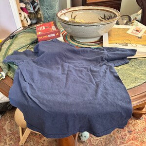 May include: A navy blue t-shirt lies on a wooden table with a green marble lamp, a ceramic bowl, and a game box labeled "Exploding Kittens".
