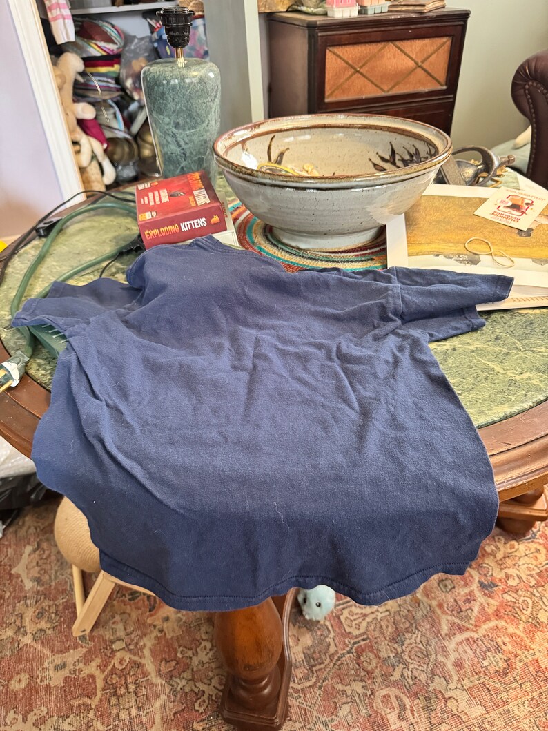 May include: A navy blue t-shirt lies on a wooden table with a green marble lamp, a ceramic bowl, and a game box labeled "Exploding Kittens".