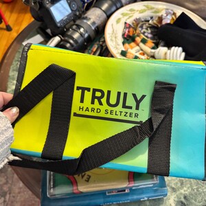 May include: A rectangular cooler bag with a gradient design of yellow, green, and blue. The bag has black straps and the words "TRULY HARD SELTZER" printed in black. The bag is held by a person.