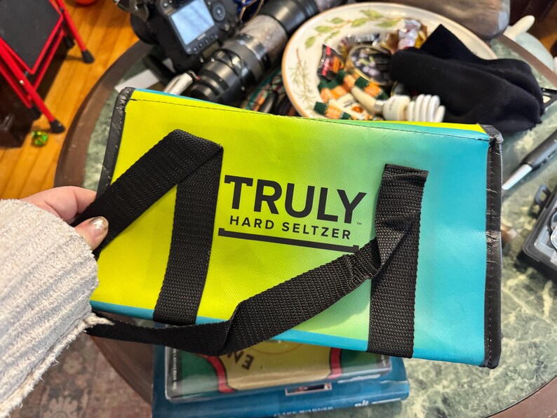 May include: A rectangular cooler bag with a gradient design of yellow, green, and blue. The bag has black straps and the words "TRULY HARD SELTZER" printed in black. The bag is held by a person.