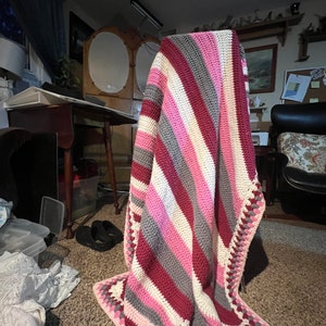 May include: A striped crocheted throw blanket in shades of pink, gray, and burgundy with a white crocheted border. The blanket is draped over a chair.