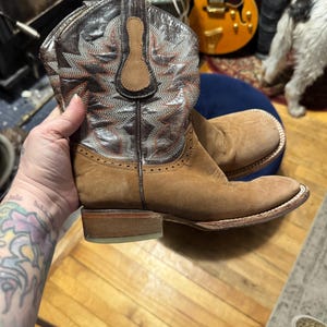 May include: A pair of brown cowboy boots. The boots have a light brown suede-like finish on the foot and a decorative stitched pattern on the upper shaft. The shaft is a darker brown with silver and orange stitching. The boots have a square toe and a low heel.
