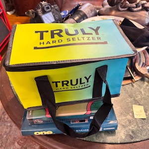 May include: A rectangular cooler bag with the text "TRULY HARD SELTZER" in black. The bag has a gradient design, transitioning from yellow to blue. It features black handles and is placed on top of books.