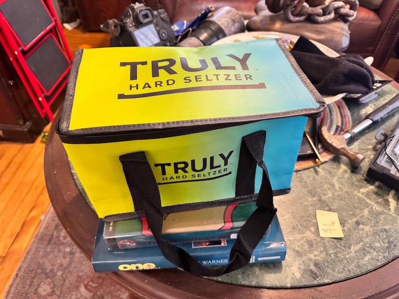 May include: A rectangular cooler bag with the text "TRULY HARD SELTZER" in black. The bag has a gradient design, transitioning from yellow to blue. It features black handles and is placed on top of books.