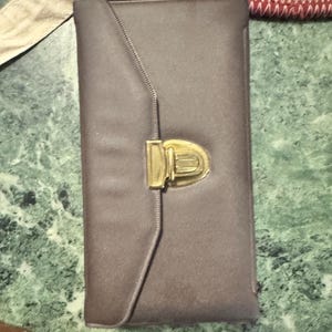 May include: A rectangular, taupe-colored clutch with a gold-toned clasp. The clutch has a flap closure and a stitched design along the edge. The clasp is a rectangular shape with a curved top. The background is a green and white marble surface.