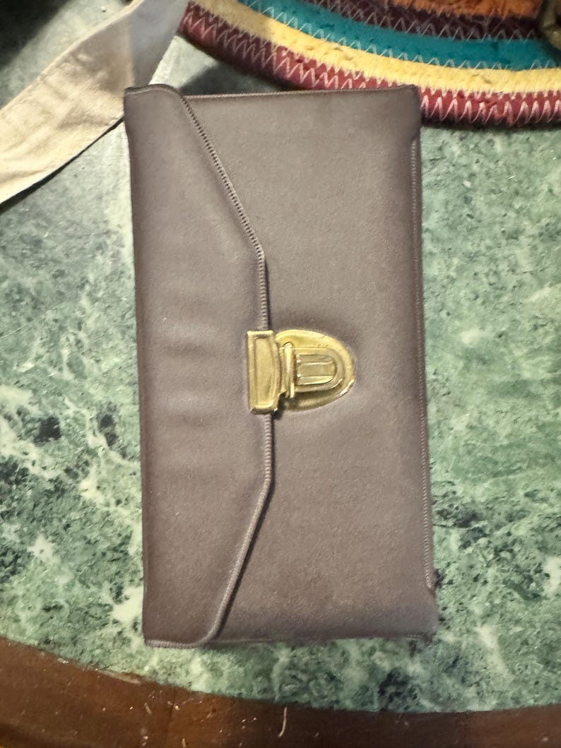 May include: A rectangular, taupe-colored clutch with a gold-toned clasp. The clutch has a flap closure and a stitched design along the edge. The clasp is a rectangular shape with a curved top. The background is a green and white marble surface.