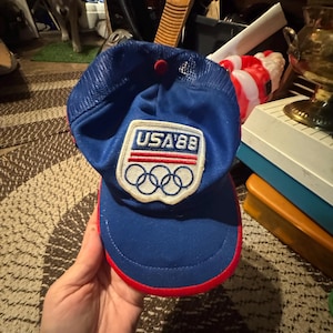 May include: A blue baseball cap with a mesh back and a red button on top. The front features a white patch with the text USA'88 above three red horizontal lines and the five interlocking Olympic rings. The brim has a red trim along the edge.