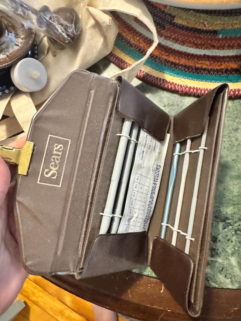 May include: A brown Sears crochet hook and needle case, open to reveal crochet hooks and knitting needles. The case has a gold clasp and the Sears logo. A chart with measurements is visible.