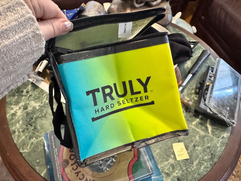 May include: A rectangular, insulated cooler bag with a gradient design of blue and yellow. The bag features the text "TRULY HARD SELTZER" in black. The bag is open, revealing a green interior.