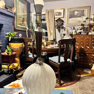 May include: A vintage table lamp with a white, ribbed glass globe base and a dark brown wooden stem. The lamp has a silver-colored metal base and accents, and a white light bulb is visible at the top. The lamp is turned on.