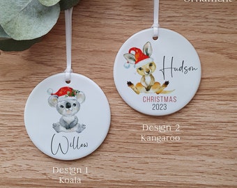May include: Two white ceramic Christmas ornaments with a koala and a kangaroo wearing Santa hats. The koala ornament says "Willow" and the kangaroo ornament says "Hudson". The ornaments are on a wooden background.