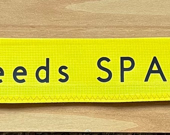 May include: Yellow fabric dog leash with black text that reads "Needs SPACE". The text is centered on the leash. The leash is sewn with a yellow thread. A blue label with white text that reads "Butta' Boo-Woo Dog Boutique" is attached to the leash.