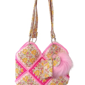 May include: A pink and white quilted handbag with a floral and character print. The bag features a pink faux fur pom-pom and a matching tag. The handles and trim are a coordinating floral pattern.