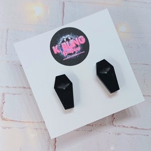 May include: A pair of black coffin-shaped earrings with a bat silhouette cutout. The earrings are displayed on a white card with the text "K BLING Boutique".