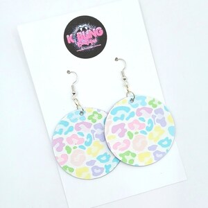 May include: A pair of pastel-coloured leopard print earrings. The earrings are round and have a white background with pink, blue, green, and yellow spots. The earrings are hanging from silver hooks.