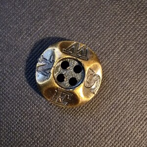 Op de afbeelding: Een gouden metalen knoop met vier gaten en de letters "M.S.W." erop gegraveerd.