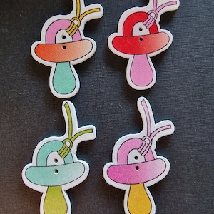 Pode incluir: Quatro botões de madeira em forma de chocalho para bebê com diferentes desenhos coloridos. Os botões são pintados em tons de rosa, vermelho, verde e amarelo. Cada botão tem um laço na parte superior.