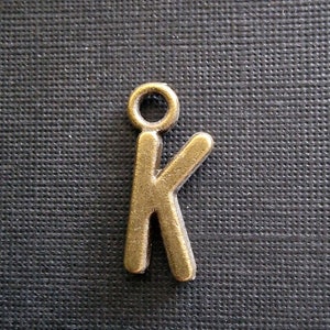 Op de afbeelding: Een gouden metalen bedel in de vorm van de letter K. De bedel heeft een lus aan de bovenkant om aan een ketting of armband te bevestigen.
