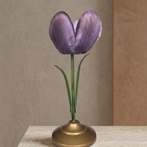 Peut inclure: Une tulipe violette unique avec une tige et des feuilles vertes, présentée dans un vase doré. La fleur est placée sur un fond neutre, créant une esthétique minimaliste. Le vase a une base arrondie et un col étroit.