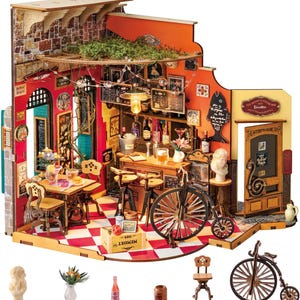 Könnte beinhalten: Miniatur-Holzmodell einer Café-Bar mit rot-weiß kariertem Boden, einer Bar, Tischen und Stühlen. Das Modell enthält ein Fahrrad, Flaschen und dekorative Elemente. Das Schild über der Tür lautet "The Artist's House Cafe Bar".