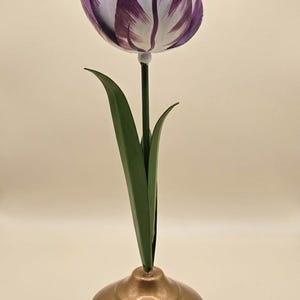 Peut inclure: Sculpture de tulipe en métal. Les pétales sont rayés de violet et de blanc, les feuilles sont vertes, et la tige est verte. Le socle rond est doré. Un élément floral décoratif pour la décoration intérieure.