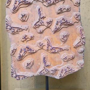 May include: A vintage wooden printing block with a butterfly pattern. The block is a warm brown color with purple butterfly designs. It is mounted on a black metal stand, showcasing the intricate details of the carved design.