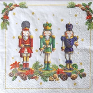 Peut inclure: Serviette en papier blanche avec trois casse-noisettes colorés en rouge, vert et bleu. La serviette est ornée de pommes de pin, d'étoiles et de verdure, créant un thème de vacances festif.