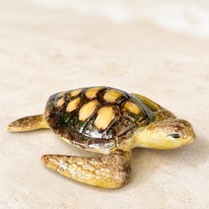 Peut inclure: Figurine de tortue de mer en céramique avec un motif de carapace détaillé dans les tons marron, jaune et vert. La tortue a un dessous jaune clair et repose sur une surface sablonneuse. L'arrière-plan est bleu clair flou.