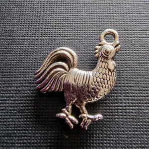 Puede incluir: Un amuleto de plata con forma de gallo. El gallo está de pie con la cabeza en alto y las plumas de la cola extendidas.