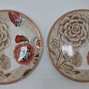 Op de afbeelding: Twee decoratieve borden met bloemmotieven. De borden hebben een beige achtergrond met grote bruine bloemen, rode en roze accenten en een gouden rand. Het bloemmotief bevat gedetailleerde bloemblaadjes en bladeren.