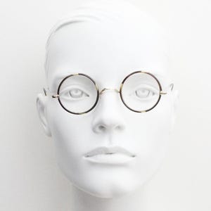 May include: Round spectacles featuring thin, metallic frames with a tortoiseshell pattern on the rims. The glasses have a simple bridge and slender temples, resting on a white mannequin head to display the circular lens shape and frame design.