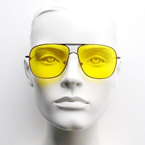 May include: Aviator sunglasses with a black frame and yellow lenses. The sunglasses feature a double bridge and a classic teardrop shape. The lenses are a bright, solid yellow, providing a striking appearance. The sunglasses are presented on a white mannequin head.