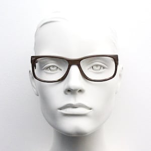 Puede incluir: Una montura de plástico marrón con lentes transparentes. Las gafas están en una cabeza de maniquí blanca.