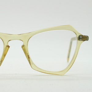 May include: Vintage spectacles with a light yellow, angular frame. The glasses have a unique shape with a pointed top and a slightly curved bottom. The lenses are clear, and the arms are thin and metallic.