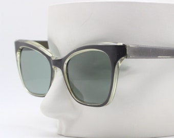 May include: Black and clear-framed sunglasses with green-tinted lenses. The sunglasses have a cat-eye shape and are displayed on a white mannequin head. The frames are a combination of black and clear plastic.