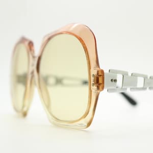 May include: Oversized sunglasses with a peach-colored frame and yellow-tinted lenses. The temples feature a silver, geometric design and black earpieces. These vintage sunglasses offer a retro aesthetic.