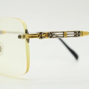 May include: Rimless spectacles with a gold and silver metal frame. The lenses have a light yellow tint. The temples feature a decorative design with gold accents. The spectacles have a classic, elegant style.