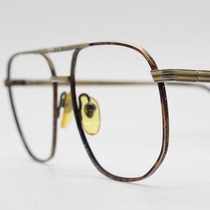 May include: Vintage aviator eyeglasses with a double bridge and a gold-tone metal frame. The lenses are clear, and the nose pads are yellow. The temples have a reddish-brown tip. A classic eyewear accessory.