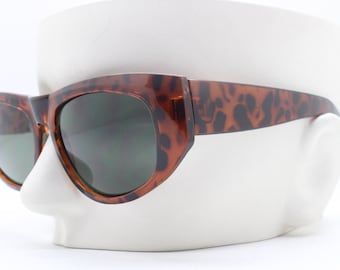 May include: Tortoise shell sunglasses with dark green lenses. The sunglasses have a cat-eye shape and are on a white mannequin head.
