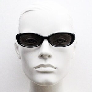 May include: Black oval sunglasses on a white mannequin head. The sunglasses have a thick frame and dark lenses, offering a sleek, classic look. The mannequin's face is smooth and pale, contrasting with the dark glasses.