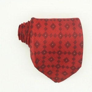 Può includere: Una cravatta rossa con un motivo geometrico ripetuto in una tonalità più scura di rosso. La cravatta è piegata e giace su una superficie bianca. Il motivo è composto da piccoli quadrati e diamanti.