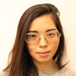 May include: A woman with long brown hair wearing glasses with a gold frame. She is wearing a grey shirt.