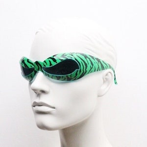 May include: Green and black zebra-striped sunglasses with a unique, curved design. The sunglasses have dark lenses and are set against a white background. The frame features a bold, eye-catching pattern.