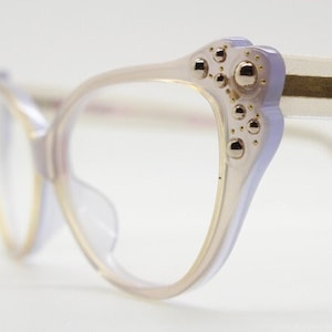 Puede incluir: Un par de gafas de ojo de gato de color morado claro con detalles dorados y tachuelas decorativas en las patillas.