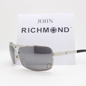 May include: Silver-toned metal sunglasses with rectangular gray lenses and black temple tips. The frame features a metallic bridge and thin arms with a small logo detail. A white card behind the glasses displays the text JOHN RICHMOND.